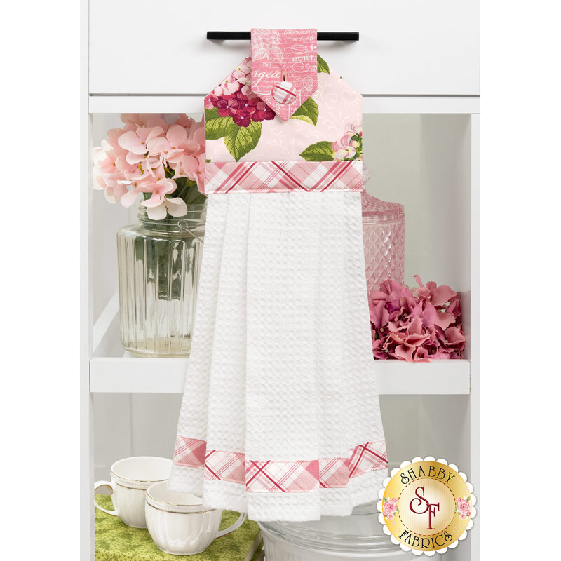 The completed Pink Hydrangeas Hanging Towel, colored in blush pink fabrics from the Hydrangeas in the Garden collection by Riley Blake Designs, hung on a white kitchen drawer and staged with coordinating flowers and kitchen utensils.