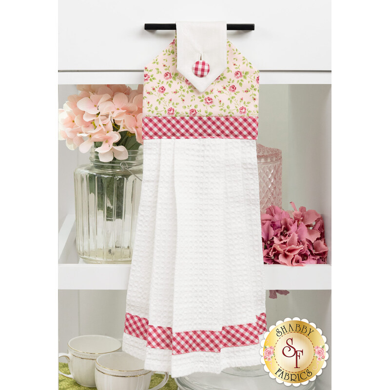 The completed Pink Roses Hanging Towel, colored in blush pink fabrics from the Hydrangeas in the Garden collection by Riley Blake Designs, hung on a white kitchen drawer and staged with coordinating flowers and kitchen utensils.