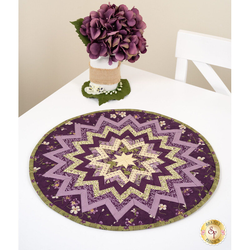 The completed Folded Star Table Topper, colored in purple, tan, and green fabrics from the Collection for a Cause - Essence series from Moda Fabrics, staged on a white table with coordinating flowers.
