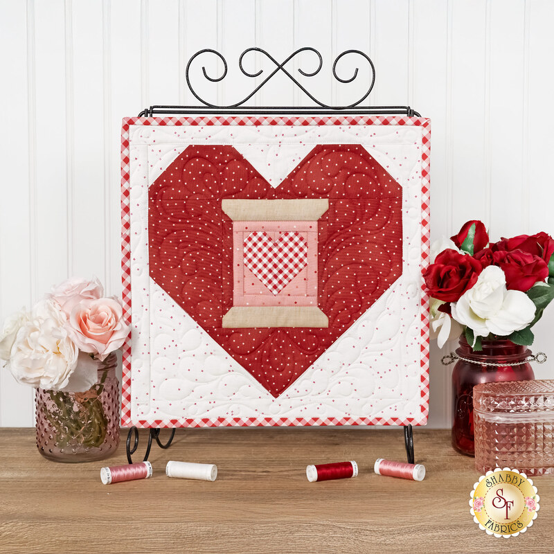 The completed February spool, featuring a pink spool with a small gingham heart within a large red heart, hung on a craft holder and staged on a wooden table with coordinating spools and décor.
