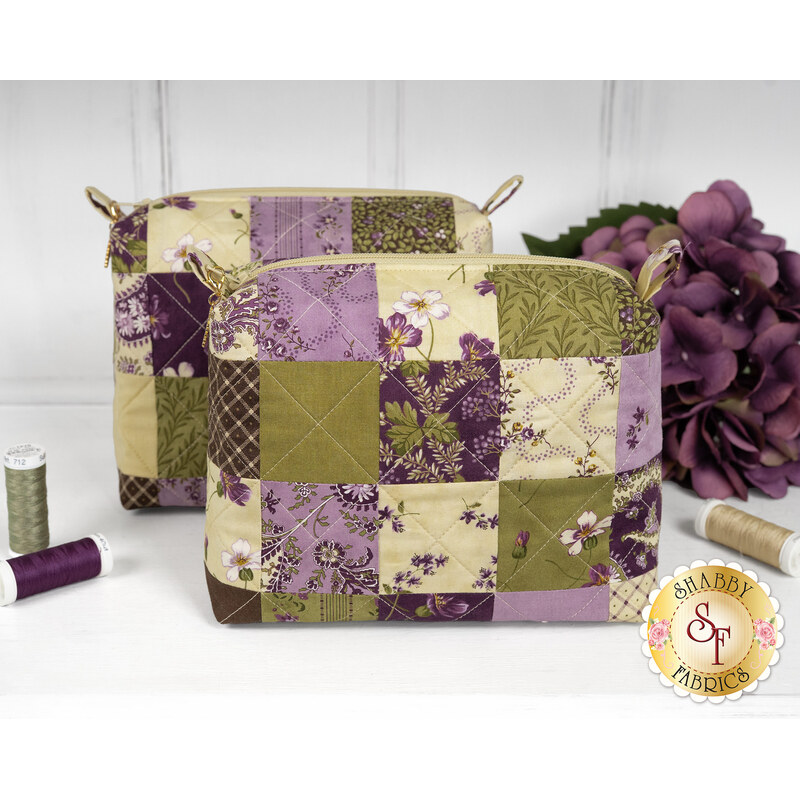 The two completed Mini-Charm Bags, colored in elegant green and purple fabrics from the Collection for a Cause: Essence collection by Moda Fabrics and staged on a white countertop, surrounded by coordinating threads and décor. A gold Shabby Fabrics logo is in the bottom right corner.