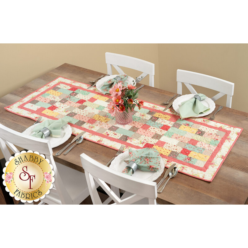 The completed Sew Charming Table Runner in lovely fabrics from the Lyda Rose collection by Moda, staged on a wooden table with four place settings and a vase of flowers in the center. 