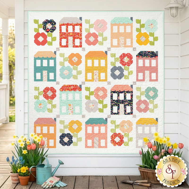 The completed Spring Town Quilt colored in vibrant fabrics from the Delightfully Yours collection by Moda Fabrics, hung on a white paneled wall on a porch with coordinating flowers and garden items.