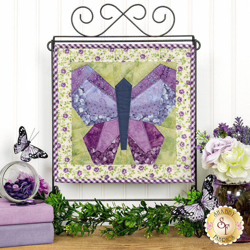 The completed April project depicting a butterfly in purple, periwinkle, green, and cream fabrics, hung on a craft display stand in front of a white paneled wall, and staged with coordinating flowers and décor.