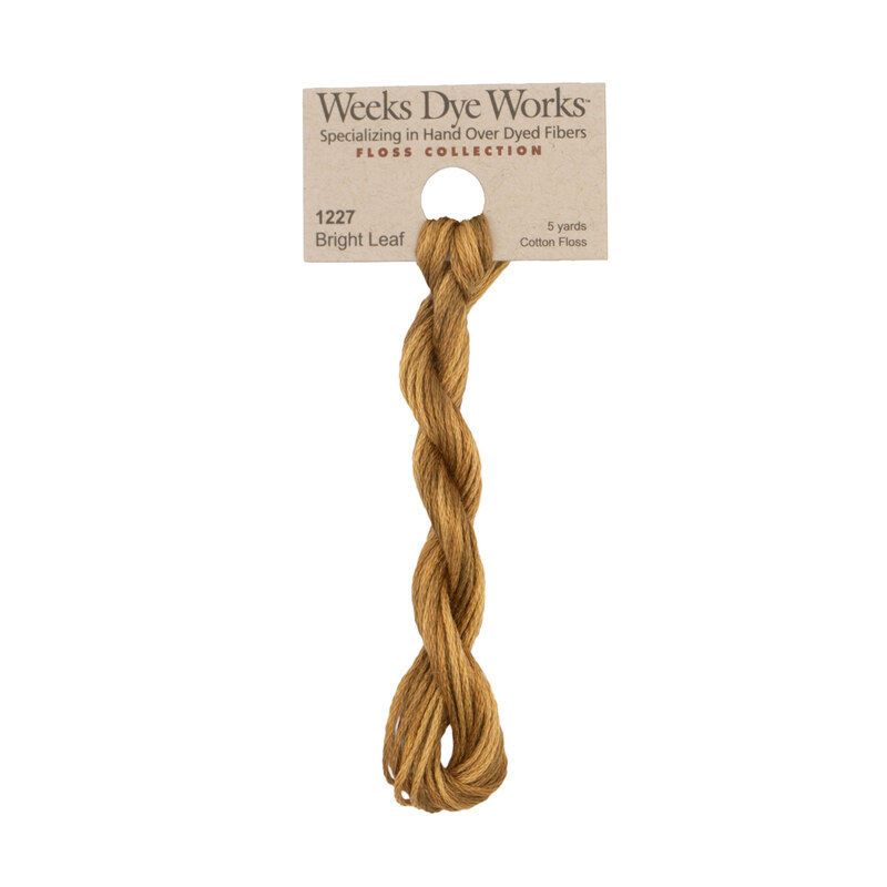 A skein of golden tan embroidery floss, twisted and bundled, with a tan label featuring the brand name and color information.