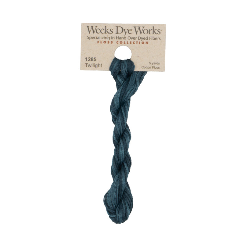 A skein of dark teal embroidery floss, twisted and bundled, with a tan label featuring the brand name and color information.