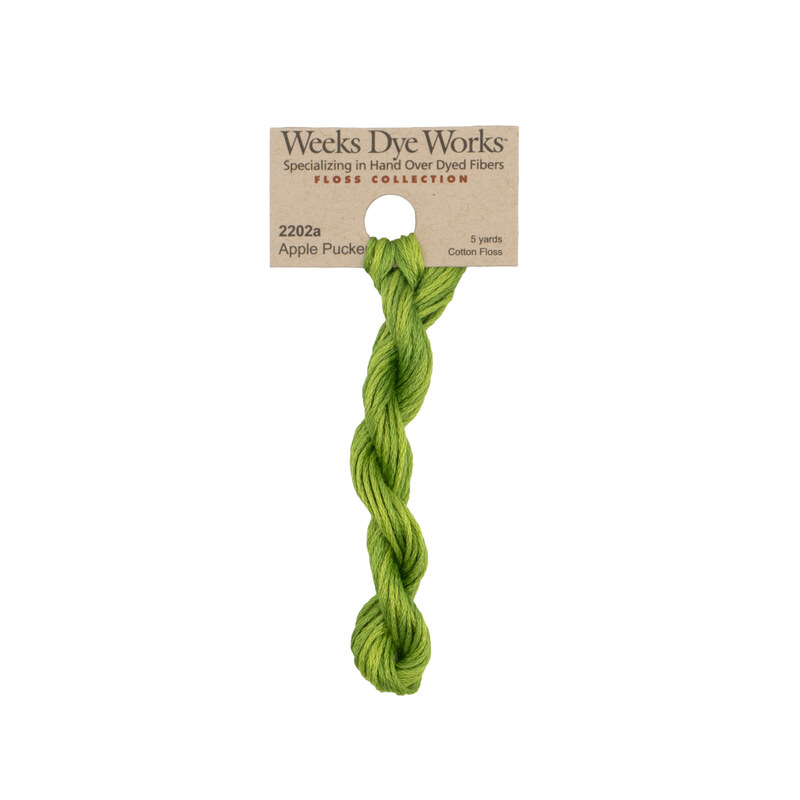 A skein of grass green embroidery floss, twisted and bundled, with a tan label featuring the brand name and color information.