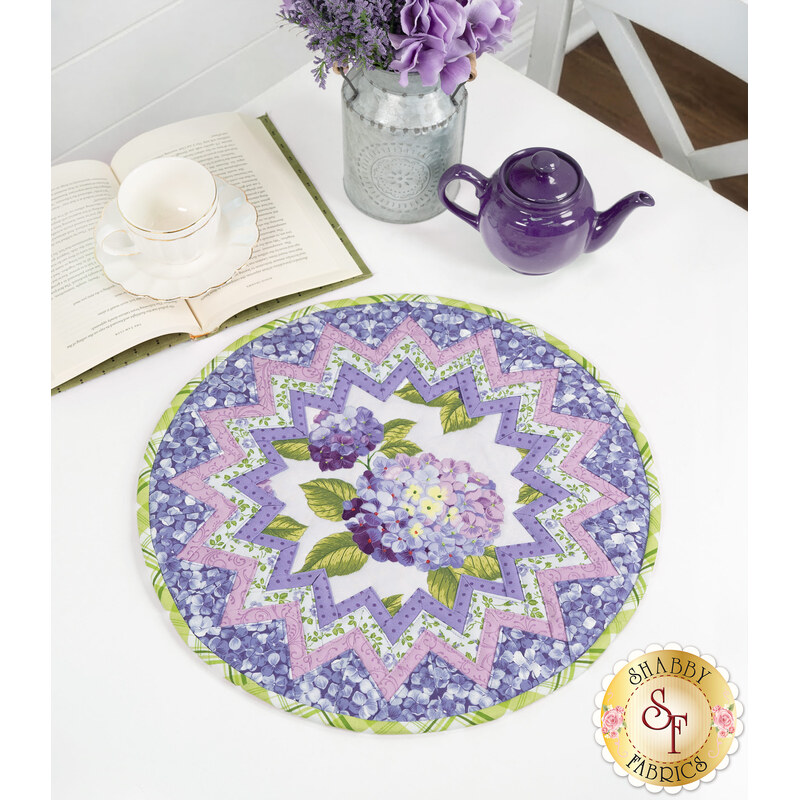 A round table topper with a bursting star design in periwinkle and lilac fabrics with blooming purple hydrangeas at the center placed on white table next to a teapot and cup and a bouquet of flowers. 