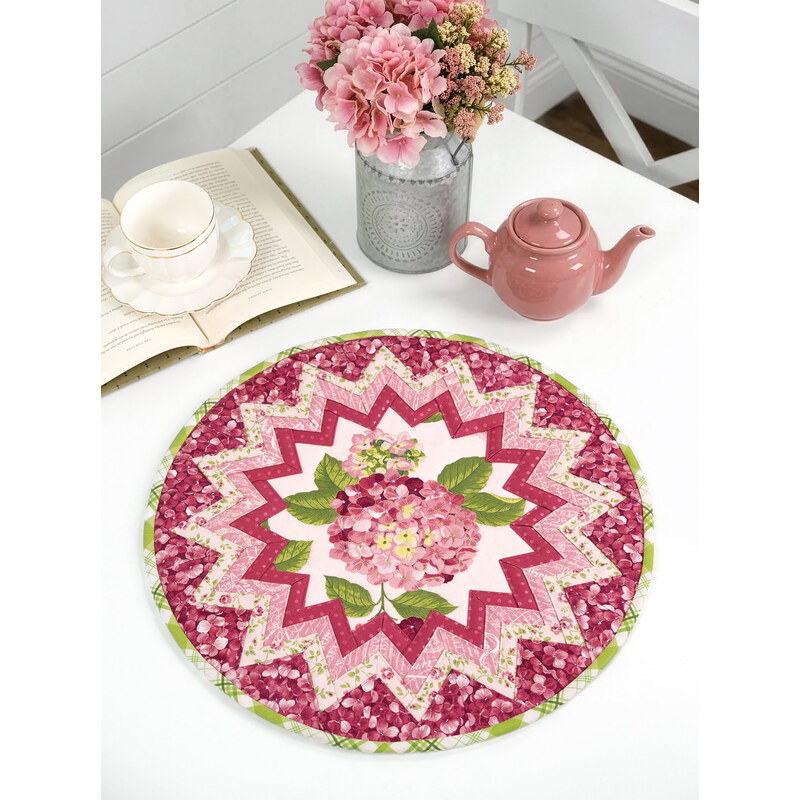 A round table topper with a bursting star design in light pink and fuchsia fabrics with blooming pink hydrangeas at the center placed on white table next to a teapot and cup and a bouquet of flowers. 