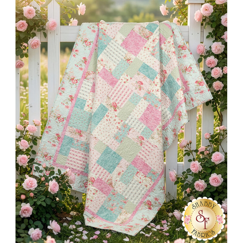 The completed Easy as ABC Quilt Kit in pastel pink, blue, and white fabrics from the Charlotte collection by Clothworks, hung over a white picket fence with pink roses.  