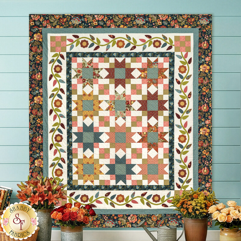 The completed Riviera Rose Quilt features a beautiful palette of teal, dusty rose, and muted autumn fabrics from the Farmer’s Daughter Collection by Andover. The design is filled with classic sawtooth stars and framed with delicate floral appliqué. It is displayed on a blue paneled wall and accented with coordinating autumn flowers.