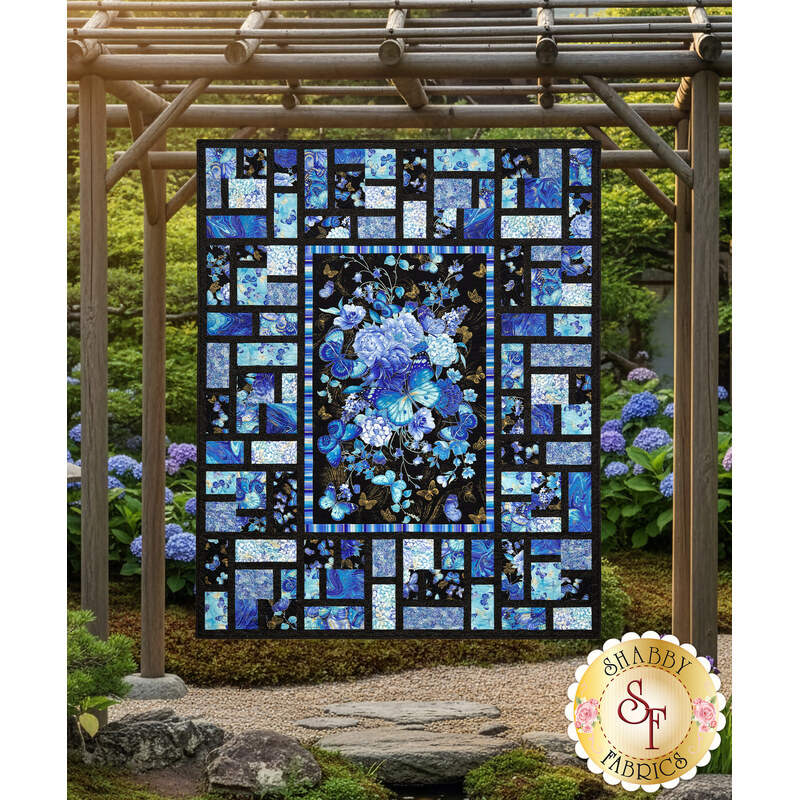 The completed Stained Glass Panel Quilt colored in the bright blue fabrics of the Escapade collection, with a stunning floral panel at the center, contrasted with striking black borders. The quilt is hanging from a wooden pergola set in a peaceful garden of blue hydrangeas with a gold Shabby Fabrics logo in the bottom right corner of the image.