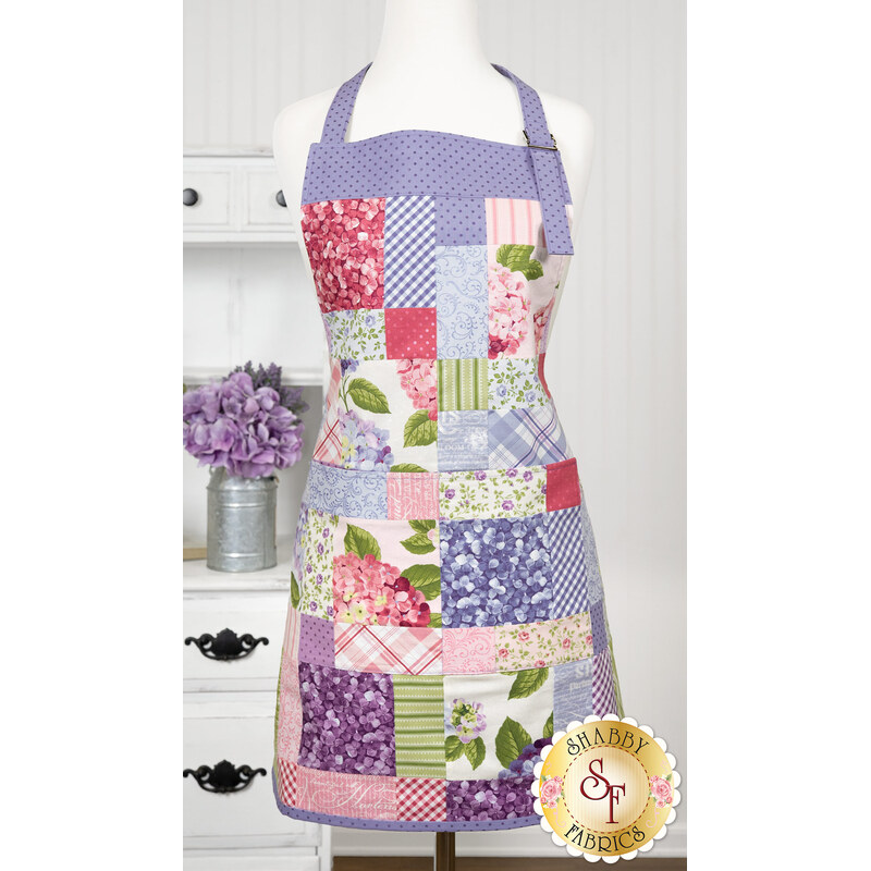 The completed Simply Charming Apron in lavender, pink, and green fabrics from the Hydrangeas in the Garden collection, displayed on a mannequin with coordinating décor in the background. 