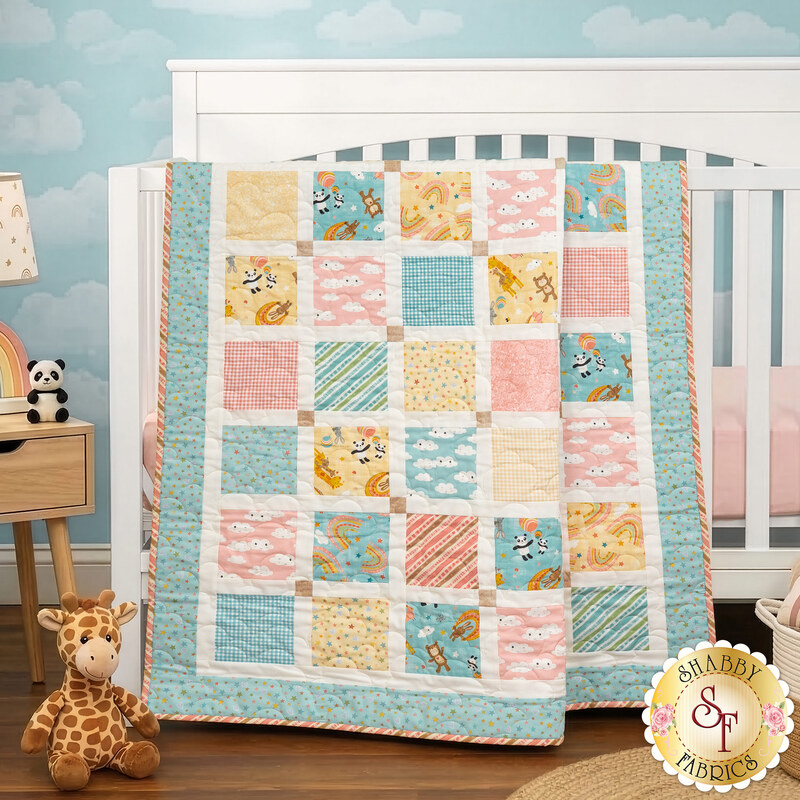 The completed Charming Windows Quilt colored in pastel primary colors from the Rainbow Dreams collection by Wilmington Prints, draped over a white crib in a nursey with coordinating décor.