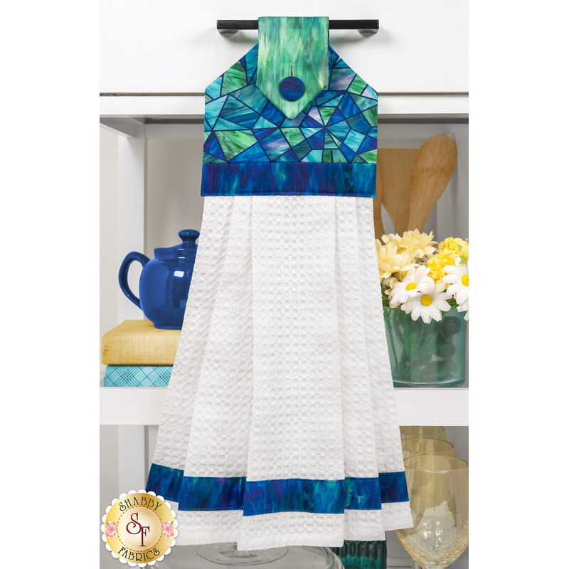 The completed Stained glass Hanging Towel, colored in blue and teal stained glass fabrics from the Risen collection by Northcott Fabrics, hung on kitchen drawer and staged with coordinating décor.