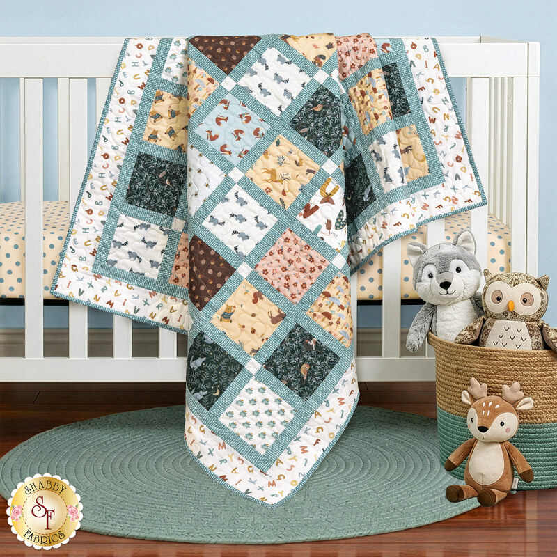 The completed Charming Windows Quilt colored in pastel pink, yellow, and blue fabrics from the Alphabet City collection by Dear Stella Designs, draped over a white crib surrounded by stuffed animals and a blue rug.