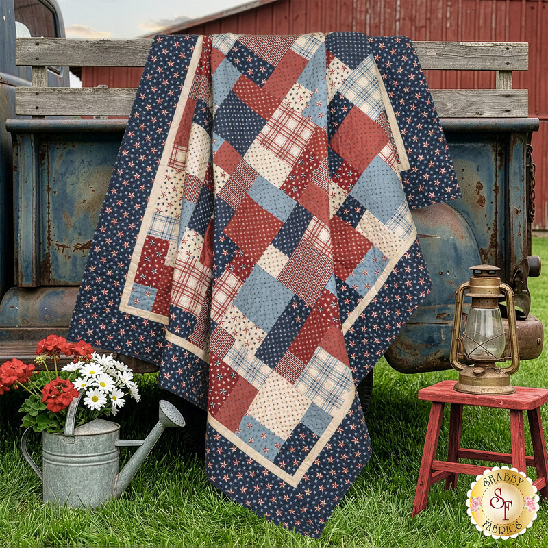 The completed Easy as ABC and 123 Quilt Kit colored in red, cream, and blue fabrics from the America 250 Years collection by Moda Fabrics, draped over an old blue truck, with a lantern and watering can nearby.
