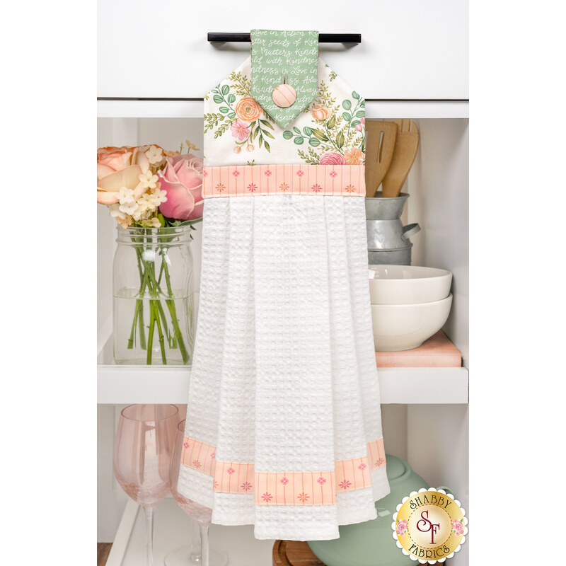 The completed Hanging Towel Precut Kit - Kindness - Cream, colored in peach, cream, and misty green fabrics from the Kindness collection by Deb Strain for Moda Fabrics, hung on a white kitchen drawer and staged with coordinating flowers and kitchen utensils.