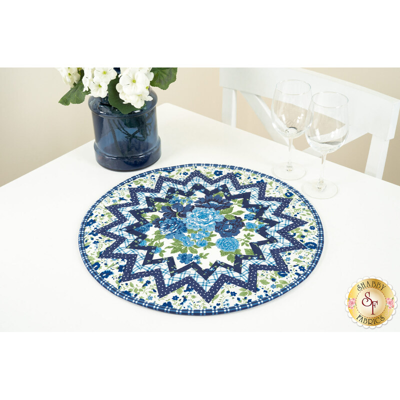 The completed Point of View Folded Star Table Topper colored in blue, light blue, green, and white fabrics from the Blueprint collection. The topper is staged on a white table with coordinating decor and tableware.