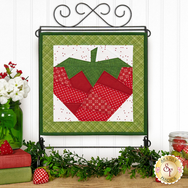  The completed June project depicting a strawberry in red, white, and green fabrics, hung on a craft display stand in front of a white paneled wall, and staged with coordinating flowers and décor.