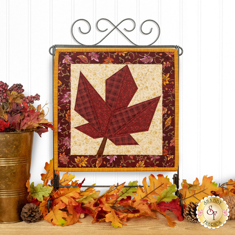 The completed Foundation Paper Piecing Series 4 November project, featuring a maple leaf in warm rust red, mahogany, and cream prints, displayed on a craft hanger and staged with coordinating foliage.