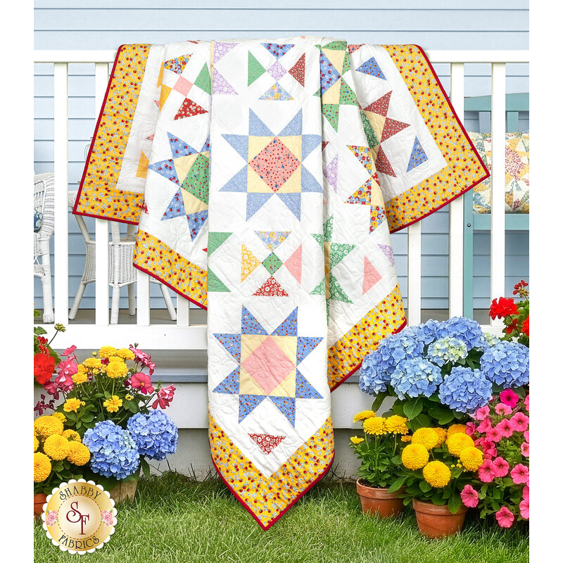 The completed vibrant patchwork Sunshine Day Quilt, colored in prints from the 30s Playtime collection by Moda Fabrics, draped on a white porch railing, surrounded by colorful flowers and a blue house.