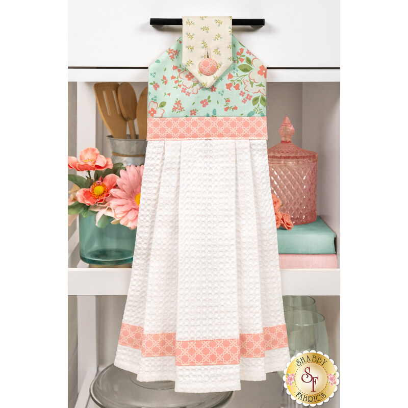 The completed Sunbonnet Hanging Towel, colored in pastel teal, soft cream, and pink fabrics from the Sunbonnet collection from Moda Fabrics, hung on a white kitchen drawer and staged with coordinating pink flowers and kitchen utensils.