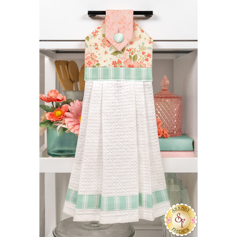 The completed Sunbonnet Hanging Towel, colored in soft cream, pastel teal, and pink fabrics from the Sunbonnet collection from Moda Fabrics, hung on a white kitchen drawer and staged with coordinating pink flowers and kitchen utensils.