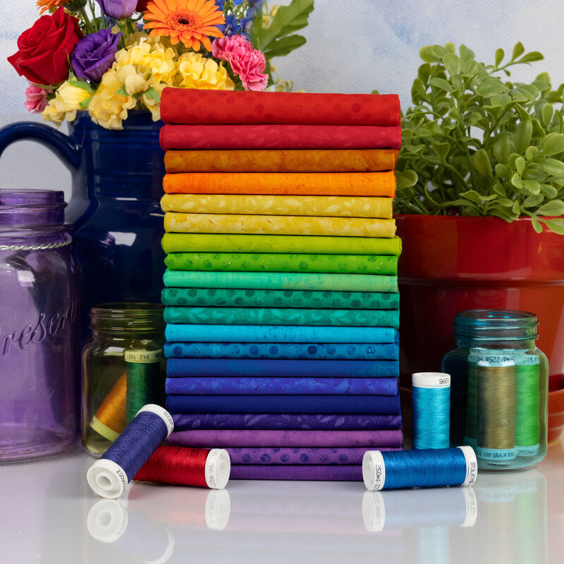 A stack of vibrant, multicolored fabric rolls arranged in rainbow order, surrounded by jars and flowers.