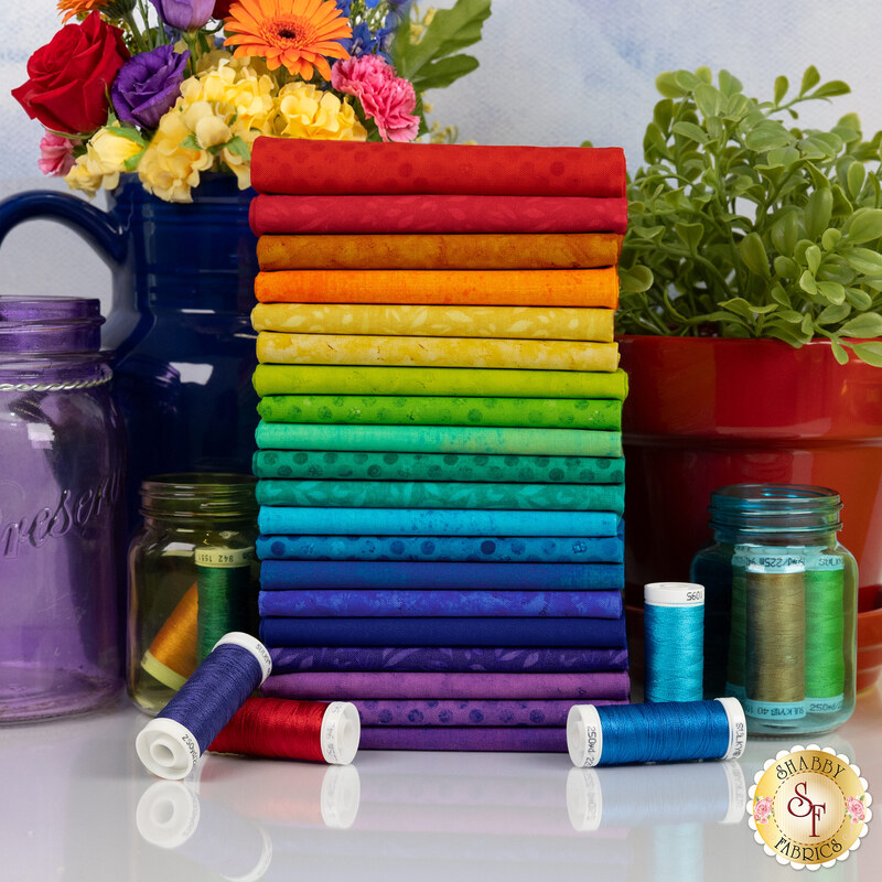 A stack of vibrant, multicolored fabric rolls arranged in rainbow order, surrounded by jars and flowers.