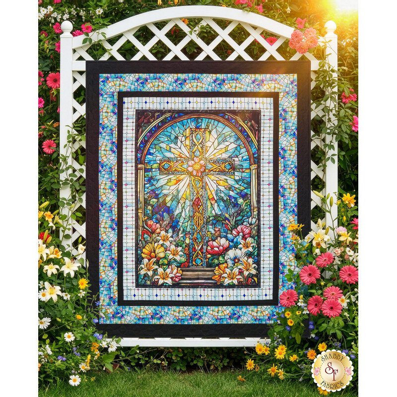 The completed Glory to God Quilt, featuring a stained-glass cross panel at its center and colorful stained-glass fabric borders from the Faith in Full Color collection by Henry Glass Fabrics. The quilt is hanging outdoors on a white trellis and surrounded by a lush floral garden, with a gold Shabby Fabrics logo is in the bottom corner.