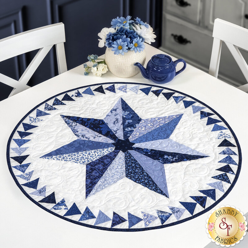 The completed Roy G. Biv table topper in blue and white fabrics from the Blue Belle collection on a white table, coordinating flowers and a tiny blue teapot sit on the corner of the table.