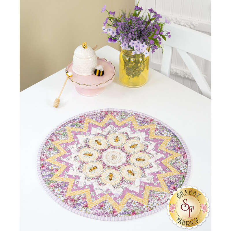 The completed Point Of View Kaleidoscope Table Topper in lavender and yellow fabrics from the Sweet Honey Bee collection by White Owl Textiles, on a white table surrounded by coordinating décor.