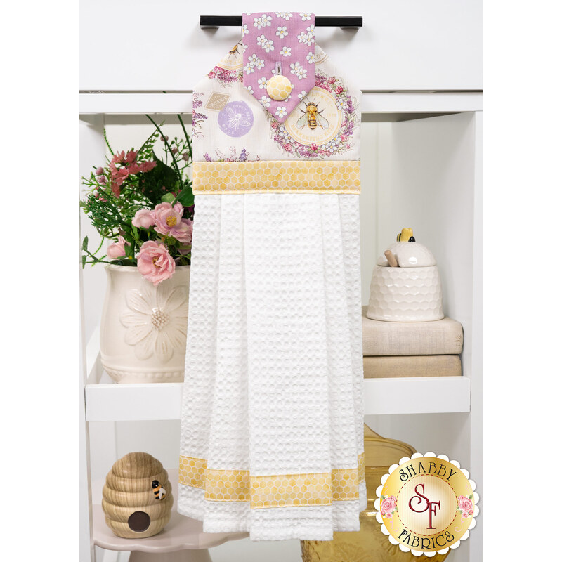 The completed Hanging Towel Kit colored in cream, yellow, and lavender fabrics from the Sweet Honey Bee collection by White Owl Textiles, hung on a drawer surrounded by coordinating décor. 