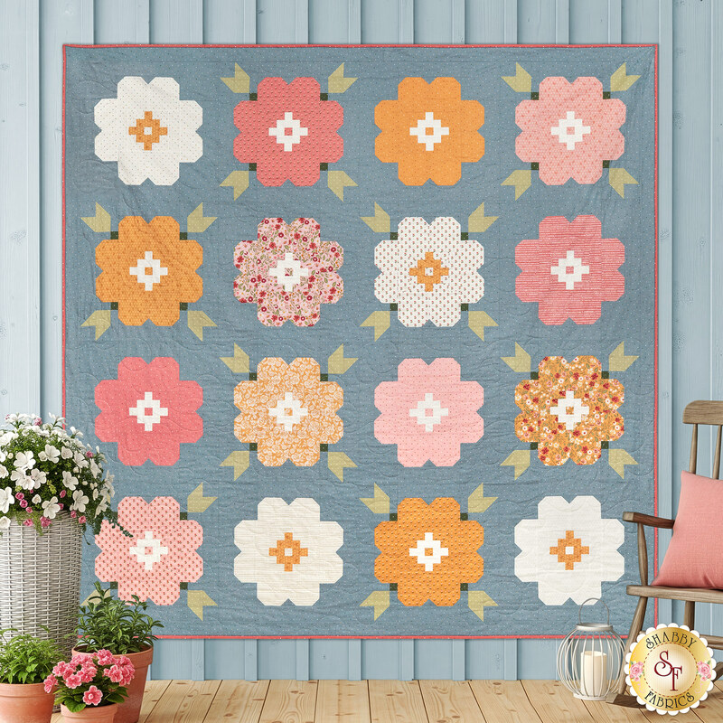 The completed Vintage Blooms Quilt, colored in blue, pink, peach, and soft white fabrics from the Wild Honey collection by Lella Boutique for Moda Fabrics, staged on a blue paneled wall and staged with coordinating flowers and décor. 