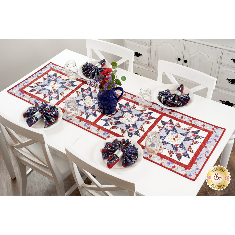 The completed table runner in red, white, and blue floral fabrics from the Forever Blooms collection, displayed on a white table surrounded by matching napkins on white plates, water glasses, and a coordinating bouquet. There is a gold Shabby Fabrics in the bottom right corner.