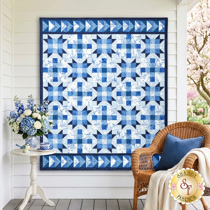 The completed Fly Over Quilt colored in blue and white fabrics from the Amalfi coast collection by White Owl Textiles. The quilt is hanging outdoors, on a white wall with coordinating blue and white décor surrounding it.