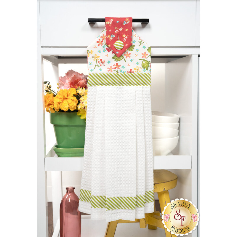 The completed Hanging Towel Kit colored in vermillion, green, and white fabrics from the In The Garden collection by Moda, hung on a drawer surrounded by coordinating décor. 