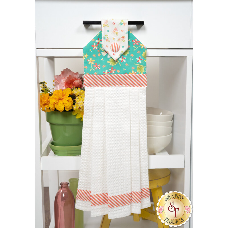 The completed Hanging Towel Kit colored in teal, orange, and white fabrics from the In The Garden collection by Moda, hung on a drawer surrounded by coordinating décor. 