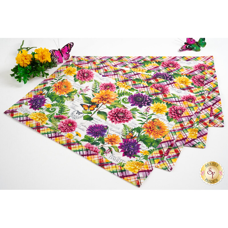 The four completed Self-Binding Placemats colored in the Dahlia Botanica collection by Wilmington Prints, fanned out and staged on a white table with coordinating flowers and butterflies.