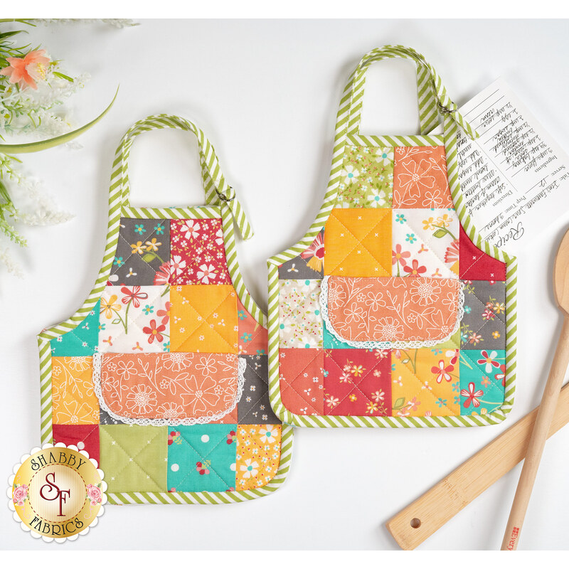 The completed Apron Hot Pads colored in red, teal, orange, and green fabrics from the In The Garden collection, staged on a white countertop with coordinating flowers and kitchen utensils; a recipe pokes out from inside one hot pad.