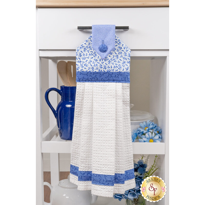 The completed Hanging Towel Kit colored in dark blue, blue, and white fabrics from the Blue Belle collection by Maywood Studio, hung on a drawer surrounded by coordinating décor. 