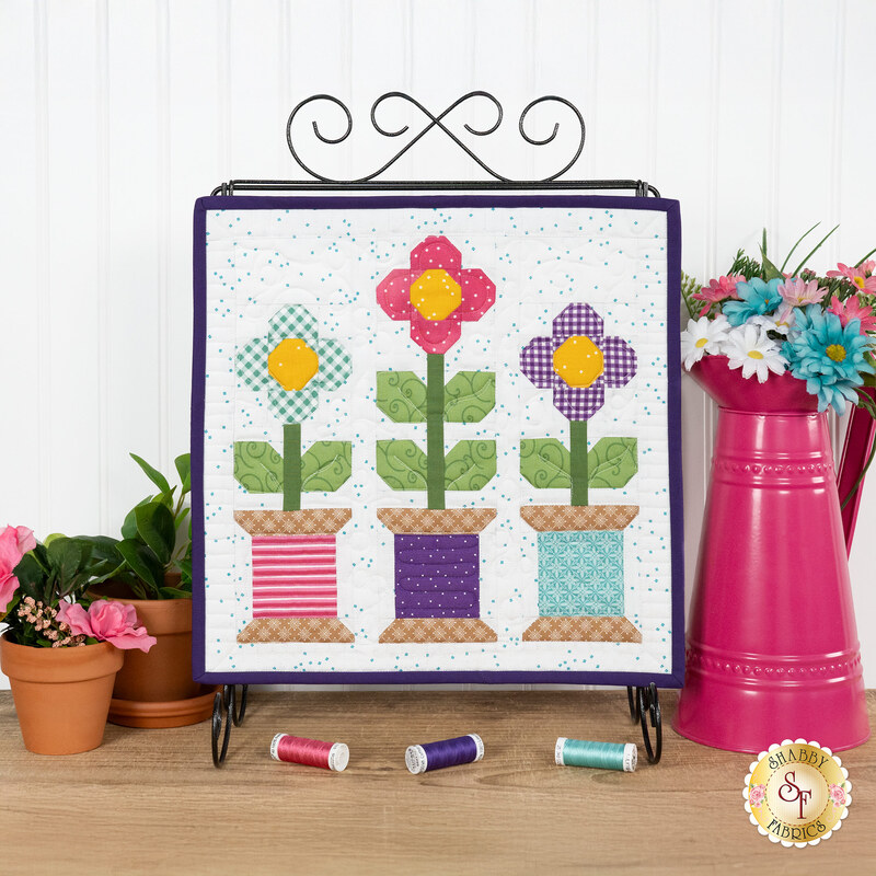 The completed May spool kit, featuring three spools with flowers growing out of them in pink, purple, and blue, displayed on a craft holder and staged on a wooden table with coordinating spools and décor.