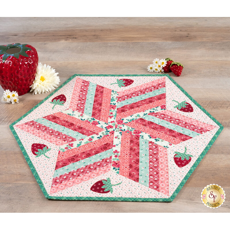 The completed 60 Degree Diamond Table Topper colored in bright pink, red, and green fabrics from the Berry & Sage collection by Riley Blake Designs, staged on a rustic wood table and paired with coordinating strawberries and daisies.
