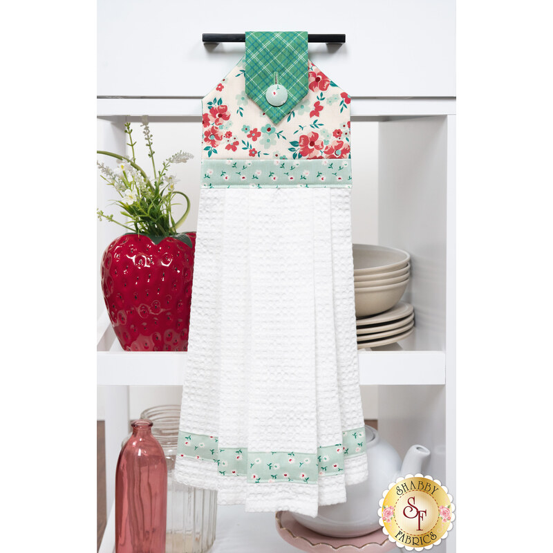 The completed Sage Hanging Towel, colored in bright green, red, and cream fabrics from the Berry & Sage collection by Riley Blake Designs, staged on a white kitchen drawer with coordinating décor and kitchenware.