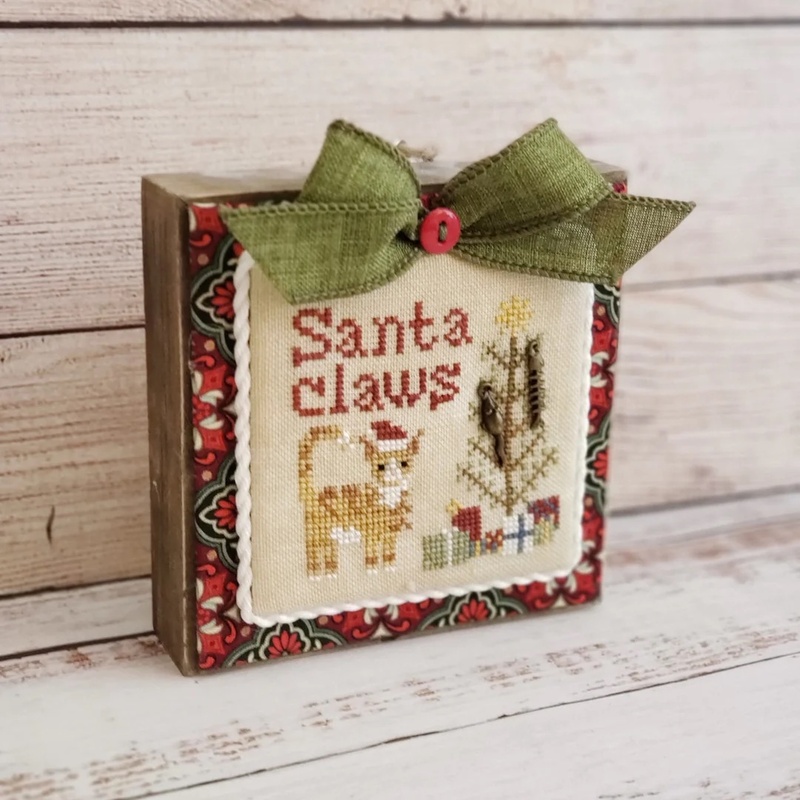 The completed Santa Claws cross stitch project, staged on a wood block with fabric and a bow.
