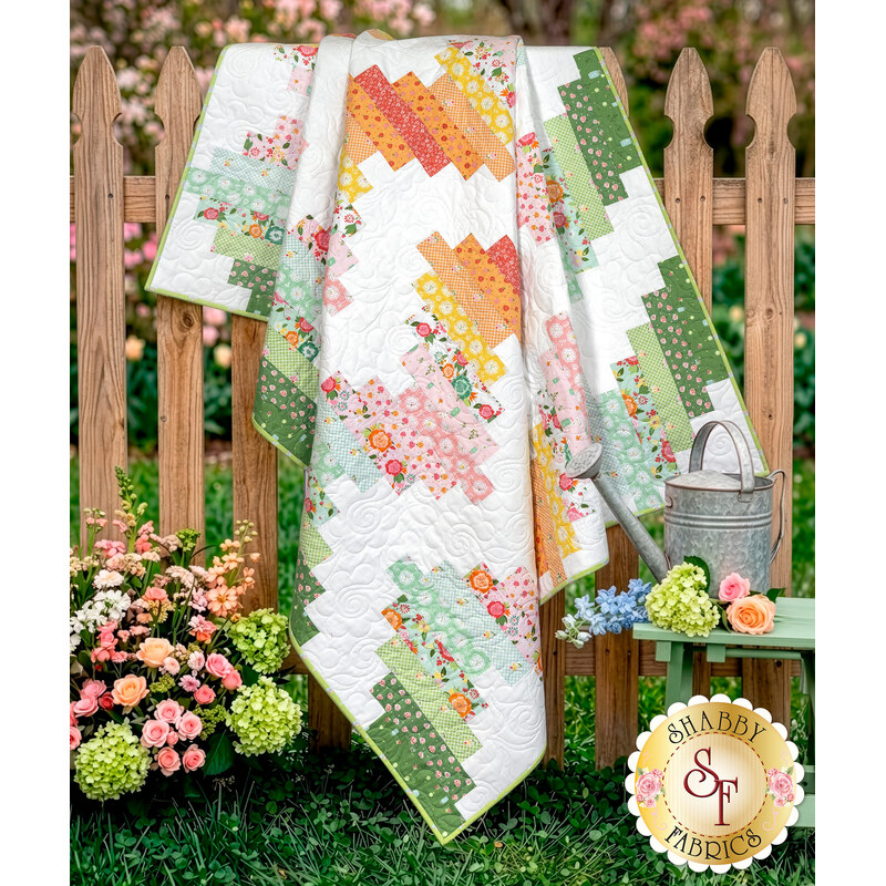 The completed Ridiculously Easy Jelly Roll Quilt colored in bright fabrics from the Summer Skies and Fireflies collection by Riley Blake Designs, artfully draped over a brown picket fence with coordinating flowers.