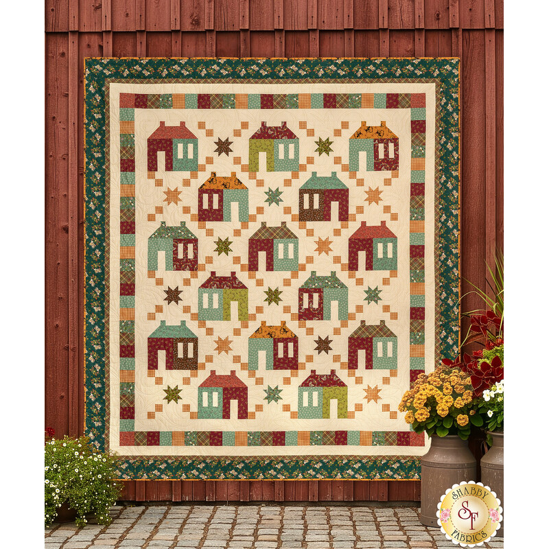 The completed Maisons de Patchwork Quilt colored in warm fabrics from the Pennyberry Farm collection by Henry Glass Fabrics, staged on a textured red barn wall with coordinating flowers and planters.