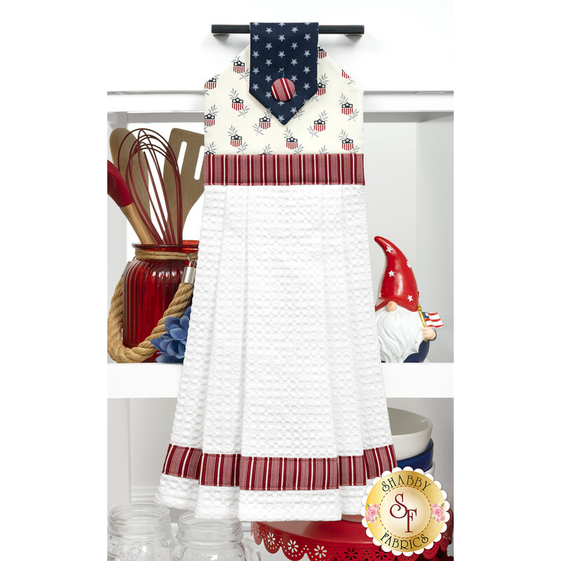 The completed Hanging Towel Kit colored in red, white, and blue fabrics from the American Gatherings III collection by Moda Fabrics, hung on a drawer surrounded by coordinating patriotic décor. 