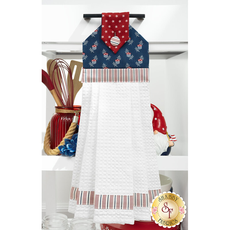 The completed Hanging Towel Kit colored in red, white, and blue fabrics from the American Gatherings III collection by Moda Fabrics, hung on a drawer surrounded by coordinating patriotic décor. 
